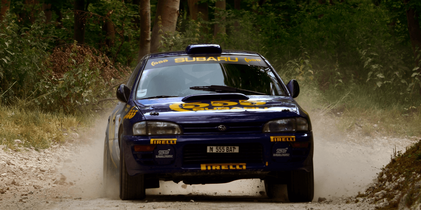 A 555-liveried Subaru Impreza rally car on a gravel forest stage, throwing up dust